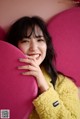 A woman in a yellow coat leaning against a pink heart shaped pillow.